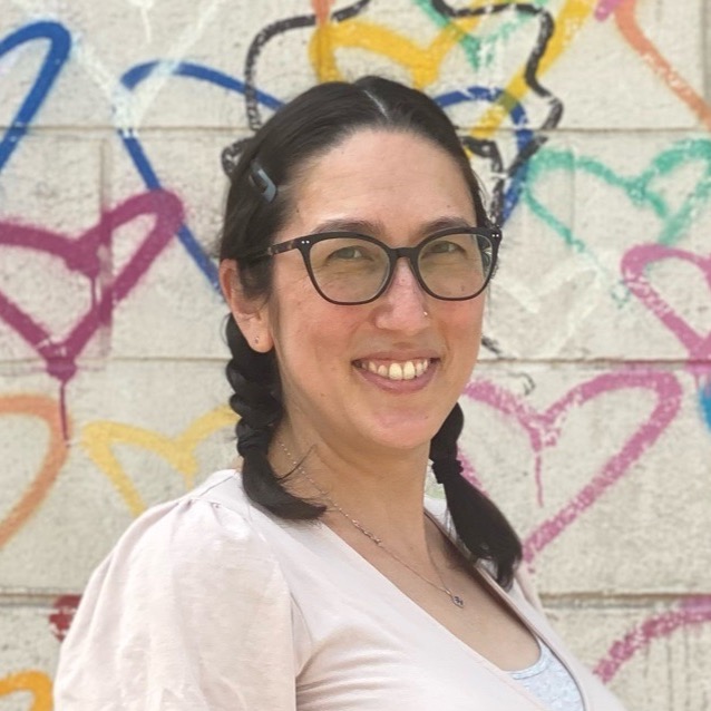 Headshot of Jess Eldredge in a circular container. She is wearing her hair in braided pigtails, wearing a pink shirt, and smiling. The backdrop is a white cement wall with colorful graffiti hearts.